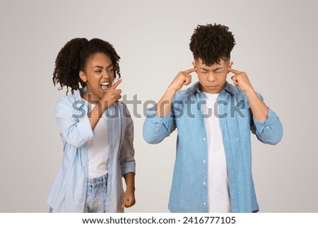 A joyful young black woman playfully teases a young man who is plugging his ears, both dressed casually in front of a neutral background, depicting a humorous interaction or mild disagreement