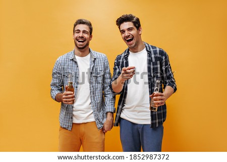 Joyful young brunet men in colorful shorts and checkered shirts laugh, point at camera and hold beer bottles on orange background.
