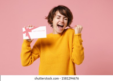 Joyful young brunette woman girl in yellow sweater posing isolated on pastel pink background studio portrait. People lifestyle concept. Mock up copy space. Hold gift certificate, doing winner gesture