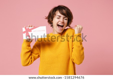 Joyful young brunette woman girl in yellow sweater posing isolated on pastel pink background studio portrait. People lifestyle concept. Mock up copy space. Hold gift certificate, doing winner gesture