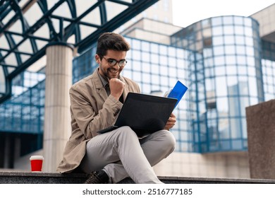 A joyful young Caucasian male entrepreneur reviews paperwork and utilizes a laptop while seated on steps outside an office building, wearing casual business attire, with a coffee cup nearby. - Powered by Shutterstock