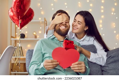 Joyful young couple celebrating St Valentine's Day. Loving woman makes surprise for her boyfriend on Saint Valentine's Day. Happy girlfriend covers man's eyes and gives him greeting card and gift box