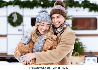 A joyful young couple embraces in snowy weather, wearing warm coats and hats, standing by a decorated camper van with holiday accents, capturing the spirit of winter romance. - Powered by Shutterstock