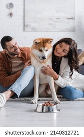 Joyful Young Couple Hugging Akita Inu Dog Near Bowl With Pet Food