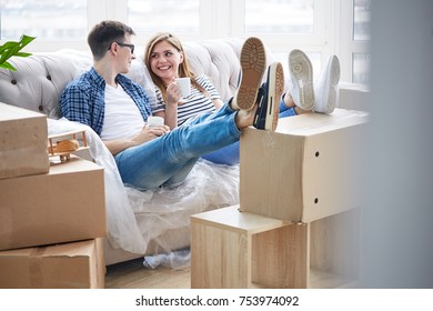 Joyful Young Couple Sitting On Sofa Covered With Plastic Wrap, Enjoying Fragrant Cappuccino And Chatting Animatedly With Each Other, Interior Of Messy New Apartment On Background