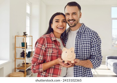 Joyful young couple stands in the living room at home, holding a piggy bank together. This portrait captures the essence of the importance of saving money and financial care within a family - Powered by Shutterstock