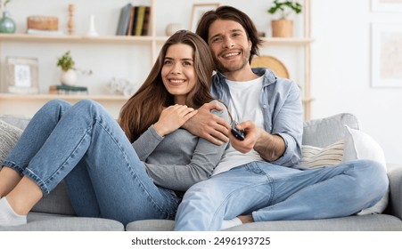 Joyful young couple watching TV together at home, embracing and sitting on couch. Cheerful man and woman switching channels on TV with remote, seeking nice movie for weekend, empty space - Powered by Shutterstock