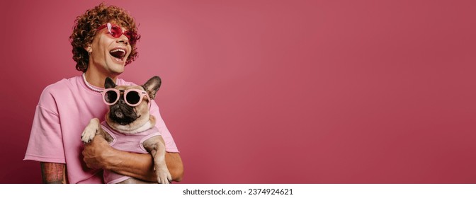 Joyful young curly hair man holding French Bulldog pet in funky glasses on pink background - Powered by Shutterstock