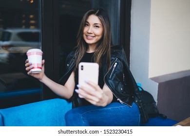 Joyful Young Ethnic Happy Female In Leather Jacket Taking Selfie With Disposable Cup Of Coffee On Mobile Phone While Sitting In Cafe And Looking At Camera