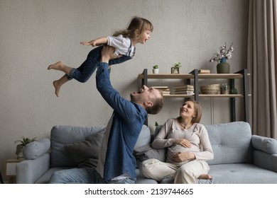 Joyful Young Father Lifting In Air Laughing Cute Small Kid Daughter While Happy Relaxed Pregnant Mother Resting On Comfortable Sofa. Cheerful Two Generations Family Having Fun On Weekend At Home.