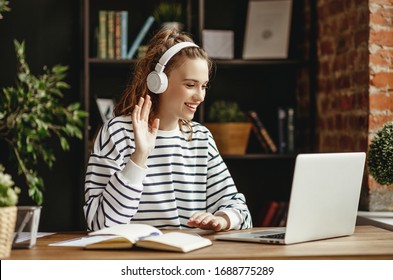 Joyful Young Female In Wireless Headphones Waving Wand Greeting To Screen While Sitting At Table And Having Video Chat With Business Partners Using Laptop Against   Of Comfortable Loft Office
