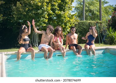 Joyful young friends sitting near swimming pool. Men and women in swimsuits and putting feet in water, smiling, giving high fives. Leisure, friendship, party concept - Powered by Shutterstock