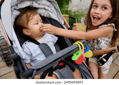 Joyful young girl feeding her baby brother with a snack while he sits in a stroller outdoors on a sunny day. Family love, sibling bond, and playful moments captured. - Powered by Shutterstock