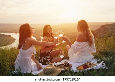 Joyful young girlfriends clinks glasses with white wine and spending time together on picnic at sunset.