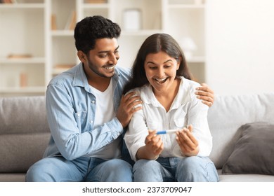 Joyful young indian couple, sitting together on comfy sofa, excitedly hold and gaze at positive pregnancy test full of hopeful smiles, in their cozy home - Powered by Shutterstock