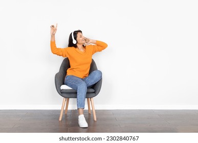 Joyful young indian woman relaxing in armchair over white empty wall, using wireless stereo headphones at home, listening to music with closed eyes and dancing, copy space - Shutterstock ID 2302840767