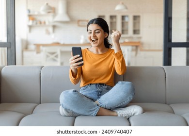 Joyful young indian woman with smartphone celebrating victory. happy excited female sitting on couch at home, holding mobile phone and shaking fist, cheering success and online win, got good news - Powered by Shutterstock