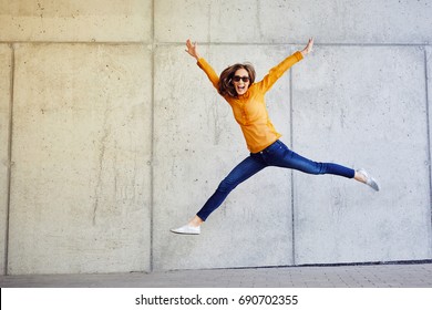 Joyful Young Lady Jumping And Raising Arms In Front Of Wall Outside