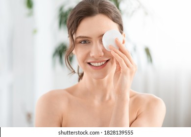 Joyful young lady removing makeup from her eyes, covering one eye with cotton pad and smiling at camera. Closeup of cheerful woman with cotton pad, using micellar water for makeup removing