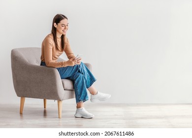Joyful young lady sitting in armchair with modern smartphone, working remotely or video chatting against white studio wall, copy space. Millennial woman watching movie, having online meeting