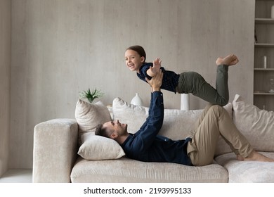 Joyful Young Latin Father Lifting In Air Laughing Cute Small Kid Daughter, Lying On Cozy Sofa At Home. Happy Two Generations Family Having Fun, Enjoying Entertaining Domestic Weekend Activity.