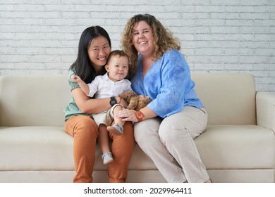 Joyful Young LGBT Family With Little Baby Boy Sitting On Sofa At Home And Looking At Camera