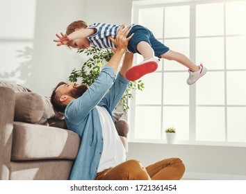 Joyful Young Man In Casual Wear Sitting On Floor And Throws Up Happy Little Son While Having Fun Together In Cozy Living Room At Home
