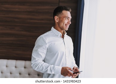 Joyful young man in classic white shirt is standing in hotel room and looking in window. He is holding cell phone in hands - Powered by Shutterstock