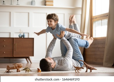 Радостный молодой человек, лежащий на ковровом полу, поднимает взволнованного счастливого маленького сына дома. В полный рост беззаботная семья двух поколений развлекается, практикуя акройогу в паре в гостиной.