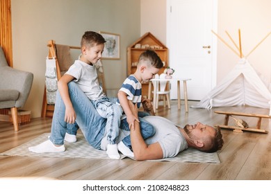 Joyful Young Man Lying On The Floor Picking Up Excited Happy Little Son At Home. Two Sons And A Father. Carefree Family Of Two Generations Having Fun. Daddy's Care And Strength.