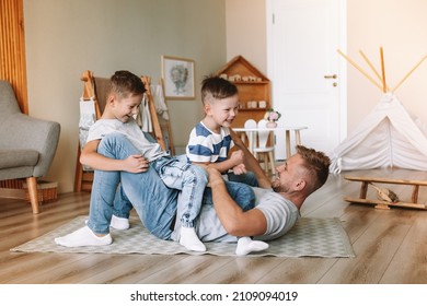 Joyful Young Man Lying On The Floor Picking Up Excited Happy Little Son At Home. Two Sons And A Father. Carefree Family Of Two Generations Having Fun. Daddy's Care And Strength.