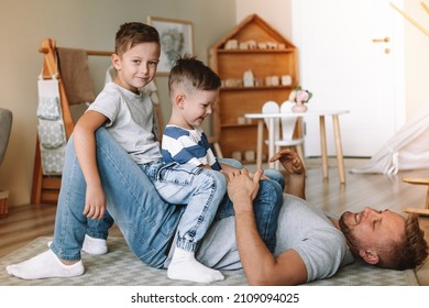 Joyful Young Man Lying On The Floor Picking Up Excited Happy Little Son At Home. Two Sons And A Father. Carefree Family Of Two Generations Having Fun. Daddy's Care And Strength.