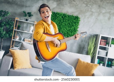 Joyful young man playing guitar indoors in modern living room filled with daylight and stylish decor - Powered by Shutterstock