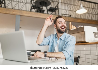 Joyful Young Man Sit At Table In Coffee Shop Cafe Restaurant Indoors Working Studying On Laptop Pc Computer Waving Greeting With Hand As Notices Someone. Freelance Mobile Office Business Concept