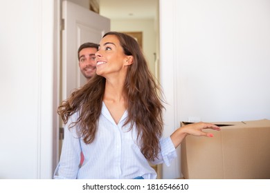 Joyful Young Man And Woman Walking Through Doorway And Looking Around In Their New Apartment With Carton Boxes. Medium Shot. New Home Or Surprise Concept