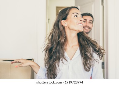 Joyful Young Man And Woman Walking Through Doorway And Looking Around In Their New Apartment With Carton Boxes. Medium Shot. New Home Or Surprise Concept