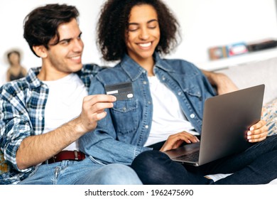 Joyful Young Mixed Race Family Couple, Guy With Girl Sitting On Sofa In Living Room, Dressed In Casual Stylish Clothes, Shopping Online Using Laptop And Credit Card, Paying For Goods, Smiling