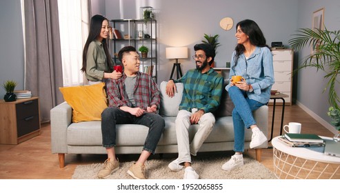 Joyful Young Mixed-races Family Couples Sitting On Sofa In Modern Living Room And Laughing In Good Mood. Asian And Hindu Men And Women At Home Drinking Coffee Or Tea And Speaking, Friends Concept