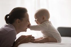 Joyful Young Mom Playing With Baby At Home, Enjoying Motherhood, Maternity Leave. Cute Infant Child Sitting On Bed, Touching Happy Mothers Face, Pushing Nose, Laughing
