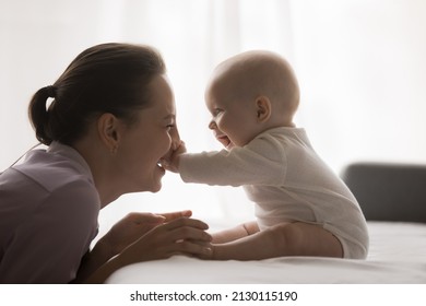 Joyful young mom playing with baby at home, enjoying motherhood, maternity leave. Cute infant child sitting on bed, touching happy mothers face, pushing nose, laughing