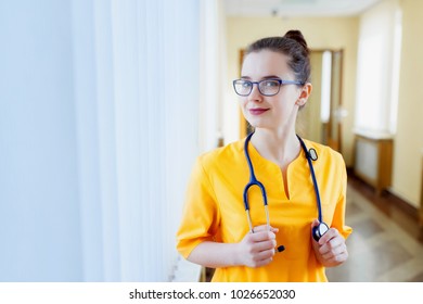 Joyful young nurse. Smiling young girl in yellow uniform, and looking at camera, in front of window clinic. The concept of health
