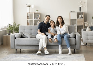 Joyful young parents and sweet kid girl sitting on soft comfortable grey couch in cozy home interior, looking at camera, laughing, smiling, promoting mortgage, property buying. Full length, wide shot - Powered by Shutterstock