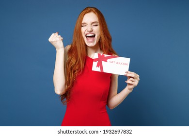 Joyful Young Redhead Woman 20s Wearing Bright Red Elegant Evening Dress Standing Hold Gift Certificate Doing Winner Gesture Keeping Eyes Closed Isolated On Blue Color Wall Background Studio Portrait