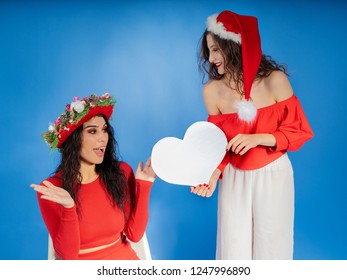 joyful young Santa girl in Christmas hat showing thumb up, holding white blank empty heart isolated on bright blue background. Happy New Year 2019 celebration holiday party concept. Mock up copy space