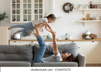 Joyful young strong woman lifting on legs arms laughing little child daughter, having fun together on cozy sofa in modern studio living room. Overjoyed small kid girl practicing acroyoga with mom.