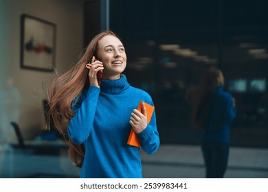 Joyful young woman chatting on the phone while holding a notebook near a glass office building on a sunny day - Powered by Shutterstock