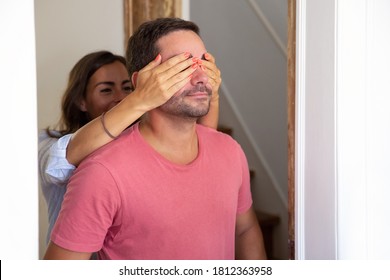 Joyful Young Woman Covering His Boyfriend Eyes With Hands And Leading Him Into Their New Apartment. Family Couple Walking Through Doorway. Medium Shot. New Home Or Surprise Concept