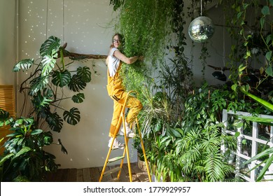 Joyful young woman gardener in orange overalls standing on a stepladder, embracing lush asparagus fern houseplant in her flower store. Greenery at home. Love of plants. Indoor cozy garden. 