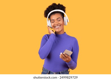 A joyful young woman is immersed in her music, smiling brightly while holding a smartphone. She wears stylish headphones and a fitted purple top, radiating happiness against a vibrant yellow backdrop. - Powered by Shutterstock