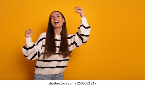 A joyful young woman with long brown hair, wearing a striped sweater, raises her fists in celebration. She smiles with excitement against a vibrant yellow background, radiating energy and happiness. - Powered by Shutterstock